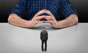 Businessman stands in front of big man sitting with his fingers clasped