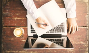 Girl writes in a notebook on a wooden desk with a cup of coffee and laptop