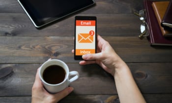 The mobile phone is in female hands. Dark brown wooden background. Diaries, tablet pc and white cup of coffee. Modern devices at home. Blank display. Woman using the touch screen.
