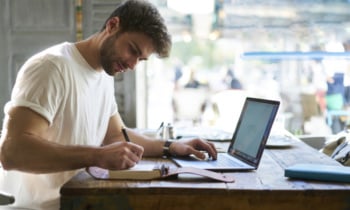 Talented hipster guy of journalism faculty of university working on modern laptop writing coursework and making notes of main thesis from text document and sitting at table near window during break