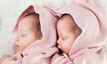 Newborn twins sisters sleeping on white fur together in cute pink sweaters