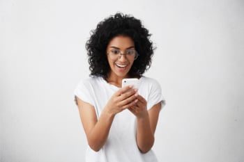 Glad attractive female with dark skin and curly black hair standing indoors with her smartphone chatting with friends. Happy cute woman in casual clothes recieving messages from her boyfriend