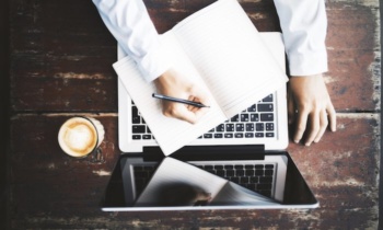 homem escrevendo em bloco de notas em frente a laptop sob mesa