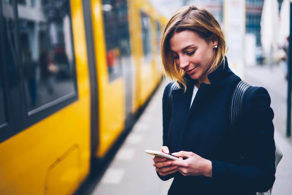 garota sorridente acessando smartphone ao lado de transporte público