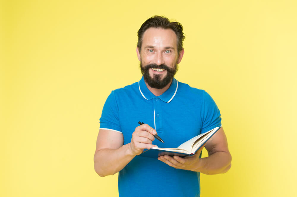homem sorridente a frente de fundo amarelo segurando caderno de anotações