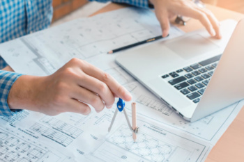 mao masculina segurando compasso em folhas de planejamentos e laptop ao lado simbolizando arquitetura da informaçao