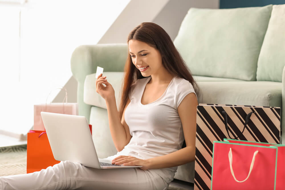 mulher sorridente sentada no chão ao lado de sacolas e fazendo compras em plataforma omnichannel em laptop