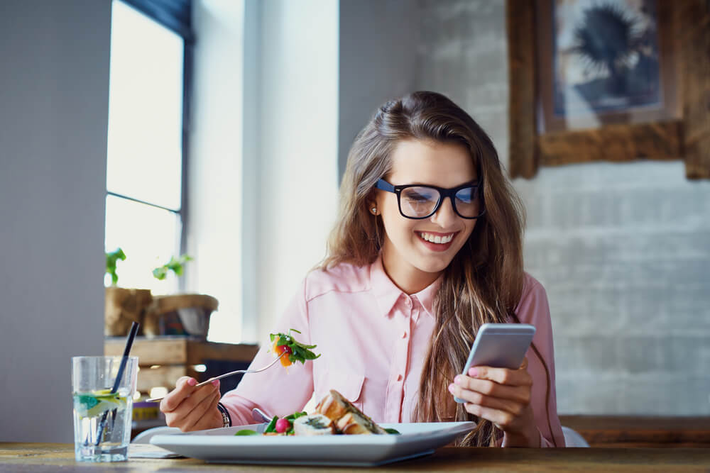 mulher sorrindo ao olhar para tela de smartphone enquanto faz uma refeiçao
