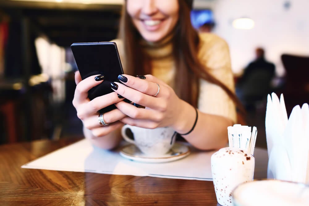 mulher em cafeteria sorrindo ao segurar smartphone com xicara de cafe em sua frente