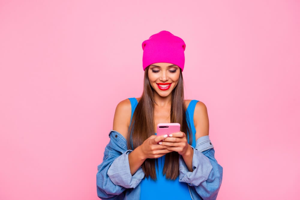 digital influencer sorrindo ao focar em smartphone em maos em fundo rosa