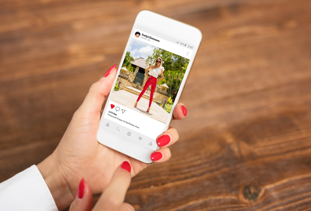mao feminina segurando smartphone sob mesa de madeira com publicaçao do aplicativo instagram em tela