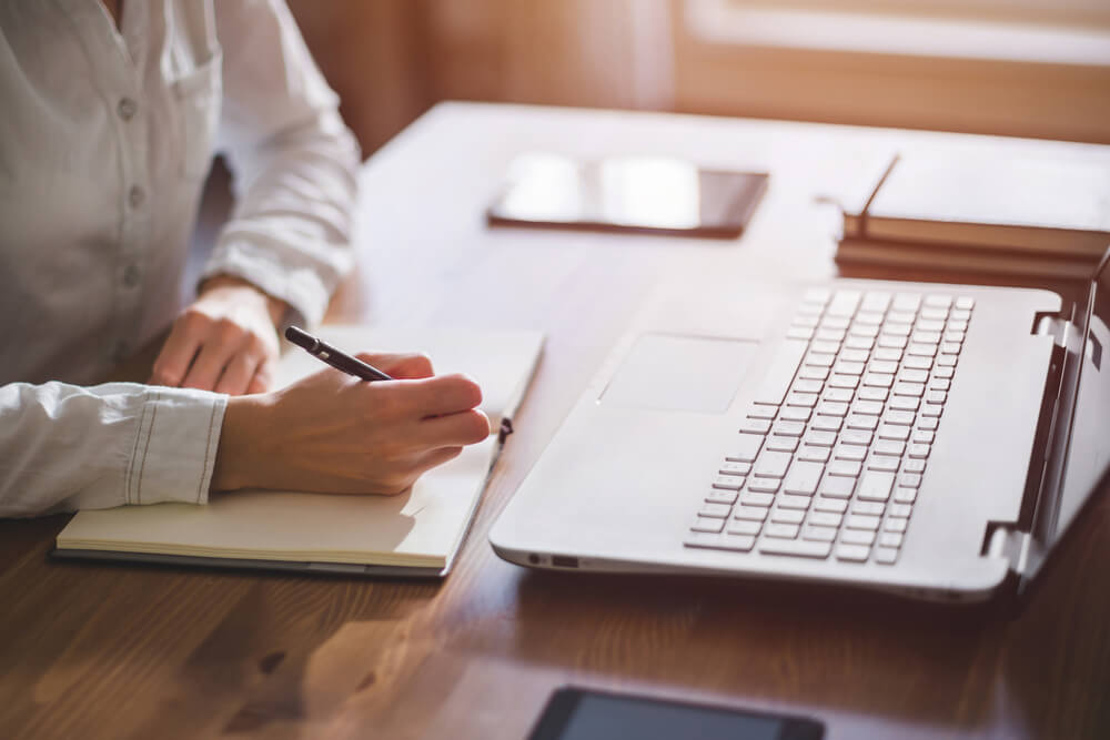 mulher escrevendo em bloco de notas em frente a laptop em mesa de escritorio