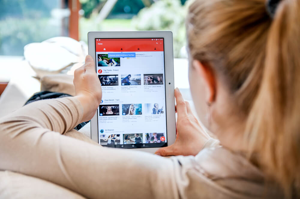 mulher sentada com tablet em maos na tela inicial do aplicativo youtube com thumbnails de videos em tela