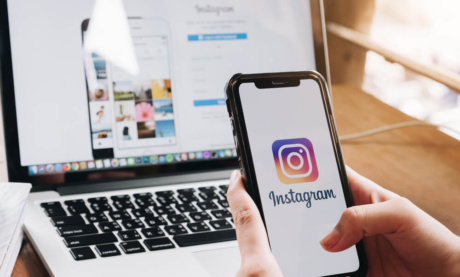 maos femininas segurando smartphone com logo de aplicativo instagram em tela em frente a laptop