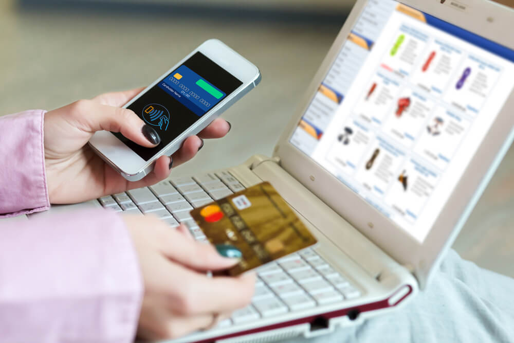 maos femininas segurando smartphone e cartao de credito em frente a laptop com pagina de loja virtual em tela