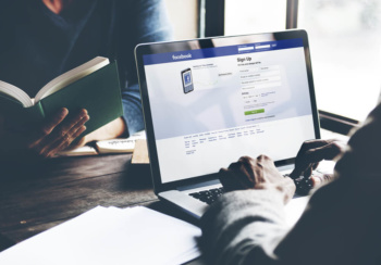 maos masculinas teclando em laptop sob mesa com plataforma facebook em tela