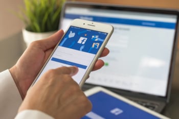 maos segurando smartphone com aplicativo facebook em tela em frente a laptop