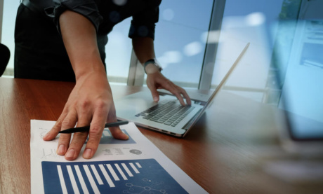 consultor em frente a mesa executiva segurando caneta e papel com graficos e com os demod em teclado de laptop