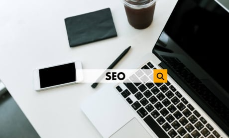 laptop em mesa com smartphone, caneta, bloco de notas e suco ao lado, com a palavra SEO em destaque