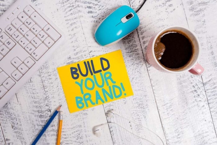 mesa de escritorio com teclado de computador, mouse e xicara de cafe ao lado de nota com a frase build your brand!