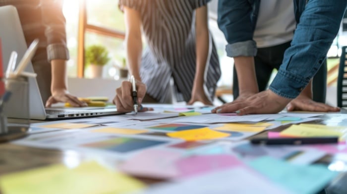 Pessoas trabalhando juntas em mesa com papéis, post-its e laptop, planejando etapas de um projeto e mostrando visualmente como fazer um briefing completo e bem estruturado.