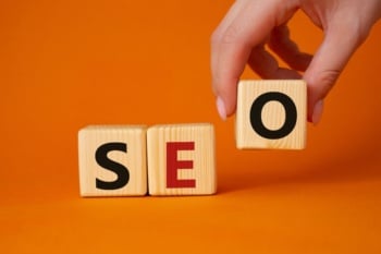 Consultoria de SEO global: mão posicionando bloco de madeira com a letra O ao lado dos blocos S e E, formando a palavra SEO sobre um fundo laranja.