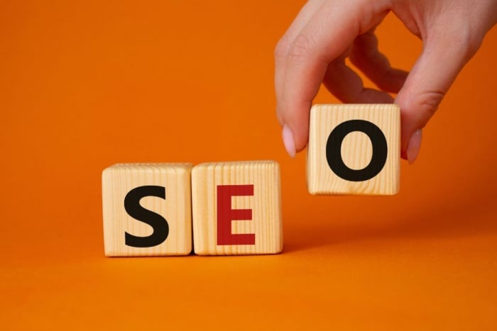 Consultoria de SEO global: mão posicionando bloco de madeira com a letra O ao lado dos blocos S e E, formando a palavra SEO sobre um fundo laranja.