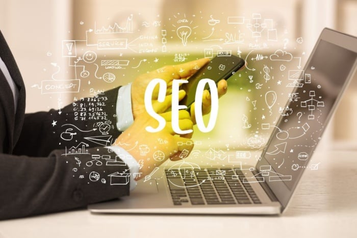 Profissional de terno de uma consultoria de SEO global, usando notebook e celular com a sigla SEO em destaque cercada por ilustrações de planejamento digital e marketing de busca.