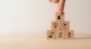Blocos de madeira empilhados em formato de pirâmide, com ícones relacionados a marketing, liderança, criatividade, relacionamento e confiança. No topo, uma mão posiciona o último bloco.