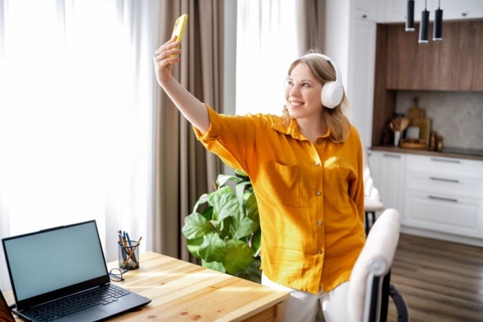 Mulher sorridente usa fones de ouvido e tira uma selfie com o celular em um ambiente doméstico iluminado, com notebook sobre a mesa ao lado.